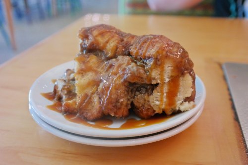 A slice of cinnamon roll topped with caramel sauce on a small white plate, resting on a wooden table.