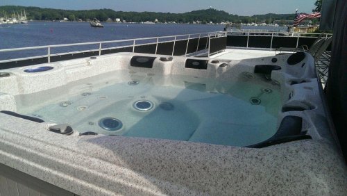 A hot tub on a deck overlooking a calm body of water, with boats in the distance and trees lining the shore.