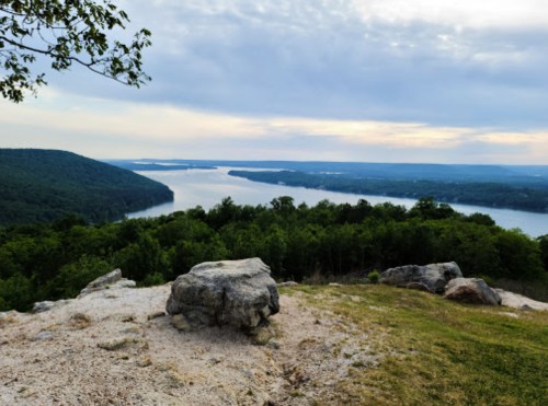 Scenic view of a river winding through green hills under a cloudy sky, with a large rock in the foreground.
