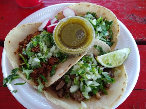 Three tacos on a plate, topped with onions, cilantro, and a side of green salsa and lime. Radishes on the side.