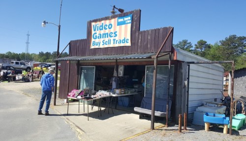 A small store with a sign reading "Video Games Buy Sell Trade," featuring outdoor tables and a clear blue sky.