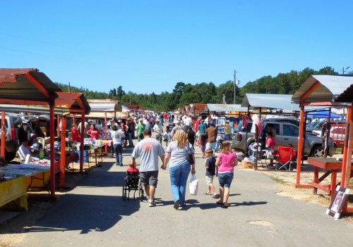 A bustling outdoor market with people walking between vendor stalls under a clear blue sky.