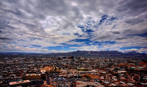 A panoramic view of a cityscape under a cloudy sky, with mountains in the distance and urban buildings below.