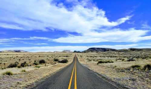 A long, empty road stretches through a vast, arid landscape under a blue sky with scattered clouds.
