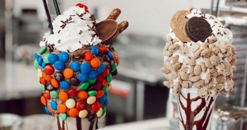 Two colorful milkshakes topped with whipped cream, candies, and cookies, one with M&M's and the other with cereal.