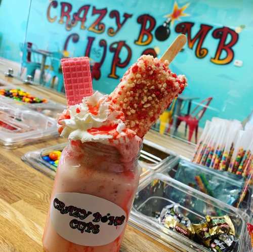 A colorful milkshake topped with a popsicle, candy, and a wafer, set against a vibrant wall with "Crazy Bomb Cup" signage.