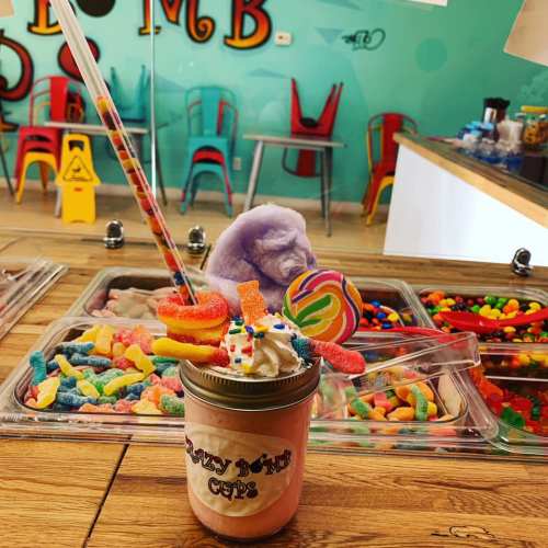 A colorful milkshake topped with whipped cream, candy, and a straw, set against a backdrop of assorted sweets.