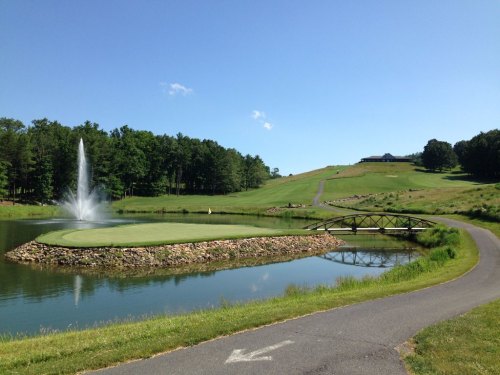 A serene golf course with a fountain, a small bridge over a pond, and lush green hills under a clear blue sky.