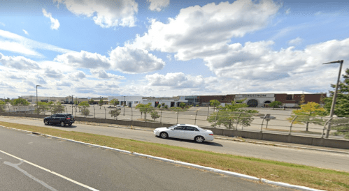 A view of a shopping area with a parking lot, cars on the road, and a cloudy sky in the background.
