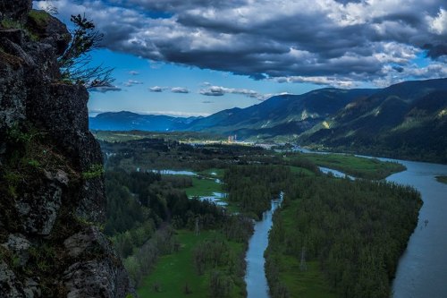 A scenic view of a river winding through lush greenery, with mountains and dramatic clouds in the background.