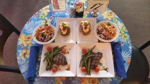 A colorful table setting with two plates of grilled steak, green beans, cherry tomatoes, and salads on a floral tablecloth.