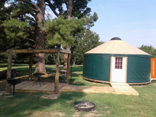 A green yurt with a tan roof beside a wooden deck and grill, surrounded by trees in a grassy area.