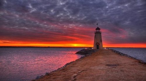 A lighthouse stands on a rocky pier at sunset, with vibrant orange and purple skies reflecting on the water.