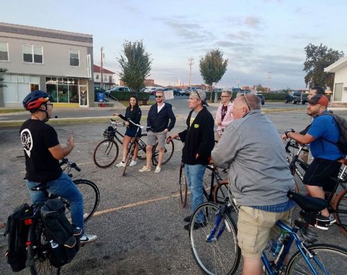 A group of people on bicycles listens to a speaker in a casual outdoor setting.