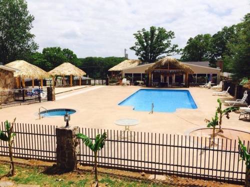 A spacious outdoor pool area with thatched-roof cabanas, lounge chairs, and a fenced perimeter surrounded by trees.