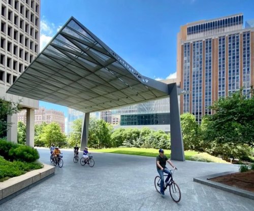 A modern pavilion in an urban park with people riding bicycles under a clear blue sky and tall buildings in the background.