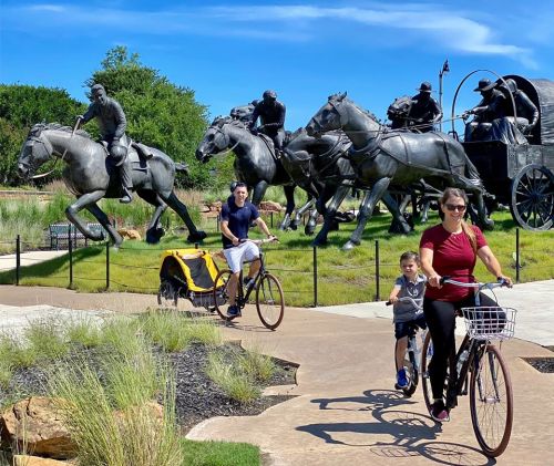 A family rides bikes past a large bronze sculpture of horses and riders in a sunny park setting.
