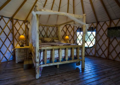 Cozy yurt interior featuring a wooden bed, bedside table, and warm lighting, with a circular window and wooden floor.
