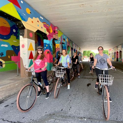 A group of women on bicycles smiles under a colorful mural in an urban setting.
