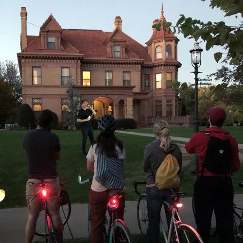 A group of cyclists watches a speaker in front of a historic mansion at dusk, with bike lights glowing.