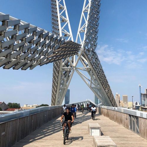 A group of cyclists and pedestrians on a modern bridge with a unique architectural design against a clear blue sky.
