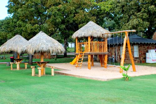Playground with a wooden playset, slide, swings, and thatched-roof shelters on a grassy area.