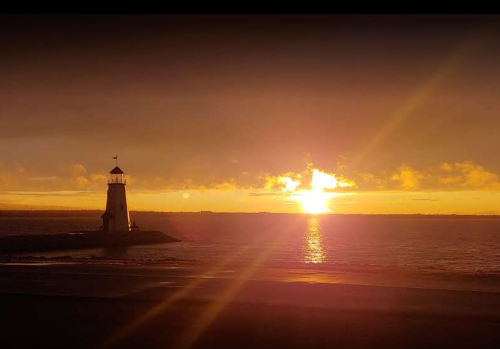 A lighthouse stands on a rocky shore as the sun sets, casting golden light over the calm water.