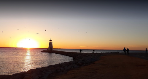 Sunset over a calm sea with a lighthouse on a rocky pier and silhouettes of people walking along the shore.