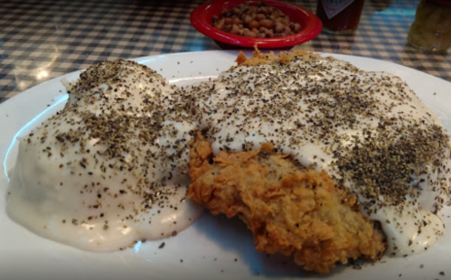 A plate of fried chicken covered in creamy gravy and black pepper, served with mashed potatoes and a side of beans.
