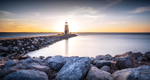 A lighthouse stands on a rocky pier at sunset, with calm waters reflecting the warm colors of the sky.