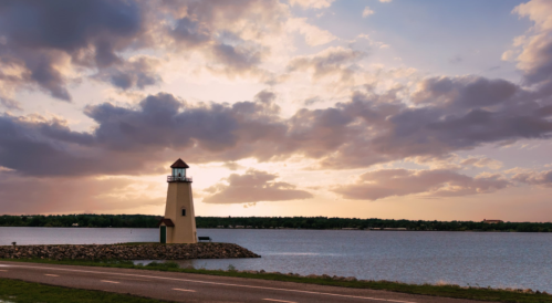A lighthouse stands by a calm lake under a dramatic sky filled with clouds at sunset.