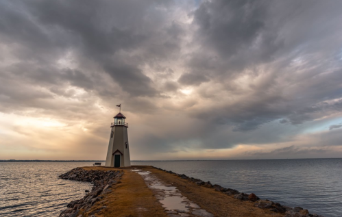 A lighthouse stands on a rocky pier under a dramatic sky, with calm waters reflecting the muted colors of sunset.
