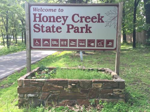 Sign welcoming visitors to Honey Creek State Park, featuring icons for amenities and activities. Greenery surrounds the sign.