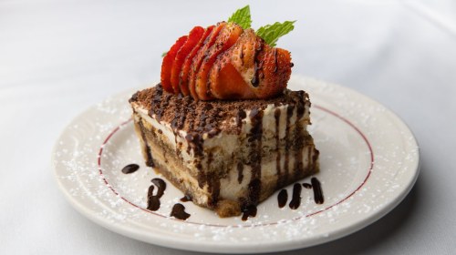 A slice of layered dessert topped with strawberries and mint, drizzled with chocolate sauce on a white plate.