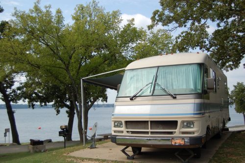 An RV parked by a lake, surrounded by trees, with an awning extended for shade.