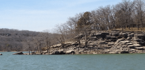 A rocky shoreline with sparse trees beside a calm lake under a clear blue sky.