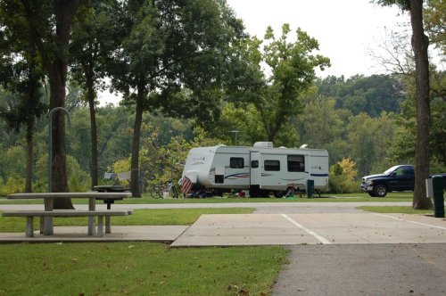 A parked RV surrounded by trees, with a picnic table and a truck nearby in a serene outdoor setting.