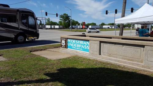 Sign for "Pedestrian Underpass Mural" near a road with an RV and a tent in the background on a sunny day.