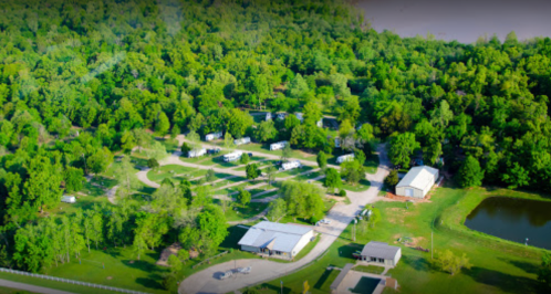 Aerial view of a green campground with RVs, buildings, and a pond surrounded by dense trees.