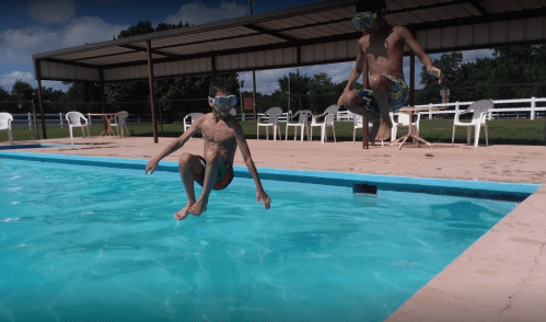 Two boys in swim trunks and goggles jumping into a pool on a sunny day, with chairs and a shelter in the background.