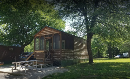 A wooden cabin with a porch, surrounded by trees and a grassy area, with a picnic table in front.