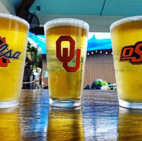Three glasses of beer on a wooden table, each featuring logos from Tulsa, Oklahoma, and Oklahoma State University.