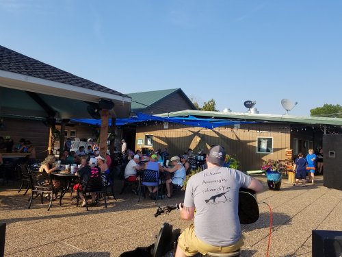 A crowd enjoys an outdoor gathering at a venue with blue umbrellas and a stage, while a musician plays in the foreground.