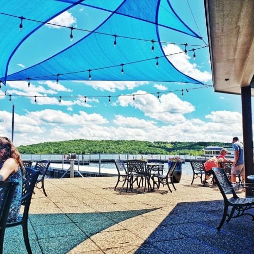 Outdoor seating area with blue shade sails, overlooking a lake and green hills under a bright blue sky with clouds.