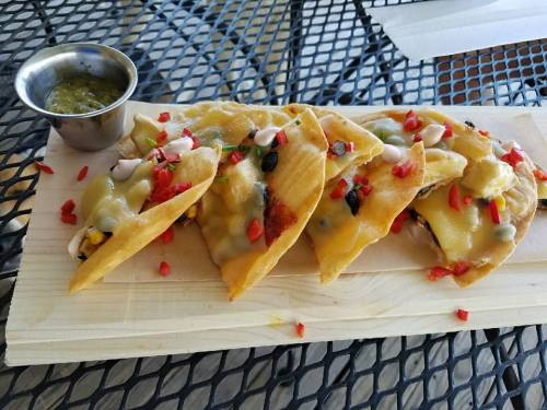 A wooden platter with cheesy tacos topped with diced vegetables and a small cup of green salsa on a metal table.