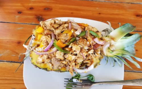 A halved pineapple filled with chicken, bell peppers, onions, and spices, served on a white plate with a fork.