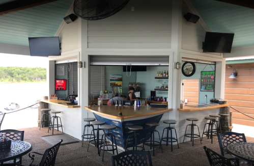 A bar with an open design, featuring a wooden counter, stools, and two TVs, surrounded by outdoor seating.
