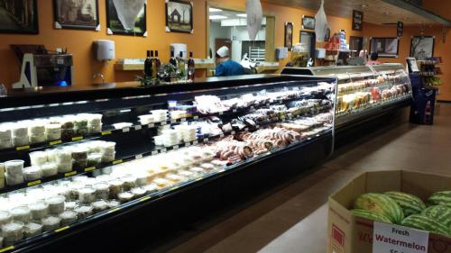 A deli display case filled with various meats, cheeses, and prepared foods, with a watermelon box in the foreground.
