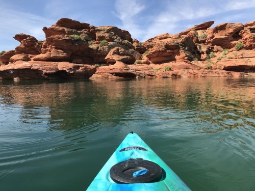 A kayak glides over calm water, with striking red rock formations and blue sky in the background.