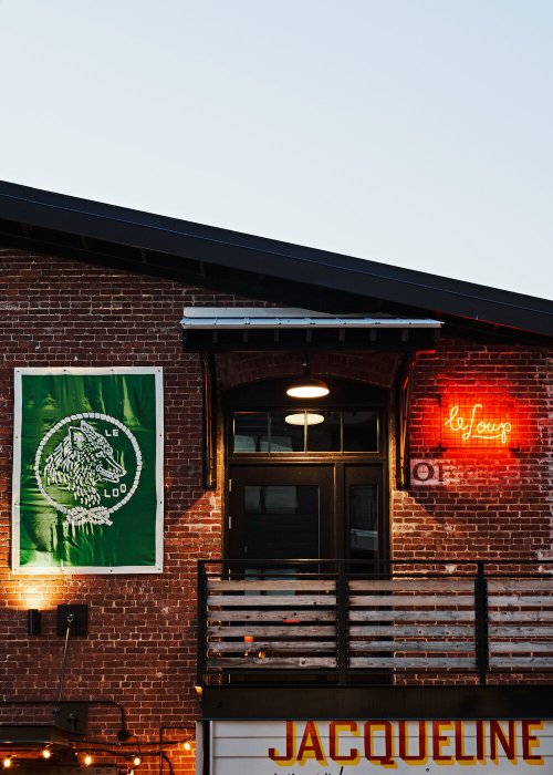 A brick building features a large green banner and a neon sign reading "le loup" against a clear sky.